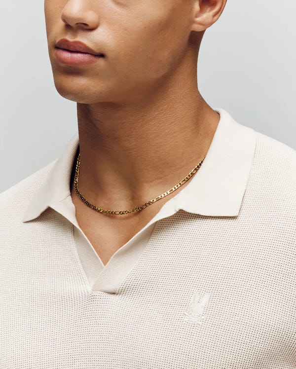 A person wearing a Psycho Bunny MENS LEETON JOHNNY COLLAR KNIT POLO in cream, made from 100% cotton with a subtle chest embroidery, paired with a thin gold chain necklace. The background is a soft, neutral tone.