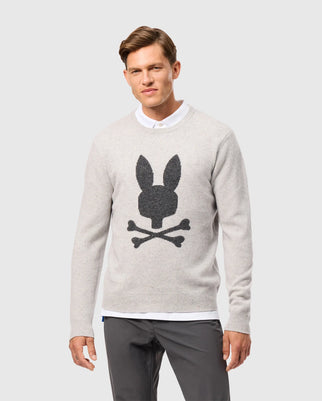 A man is wearing a light gray Psycho Bunny MENS LINCOLN MERINO WOOL SWEATER (Product Code: B6E831D200), adorned with a black skull and crossbones design that includes rabbit ears. With short hair, he has paired it with dark gray pants and a white collared shirt underneath, exuding timeless style.
