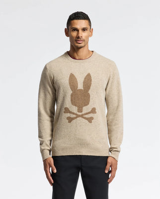 A man wearing a MENS LINCOLN MERINO WOOL SWEATER by Psycho Bunny, which features a striking large rabbit and crossbones design, stands against a plain white background.