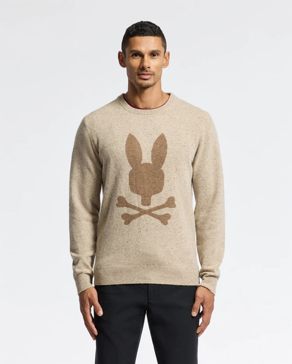 A man wearing a MENS LINCOLN MERINO WOOL SWEATER by Psycho Bunny, which features a striking large rabbit and crossbones design, stands against a plain white background.