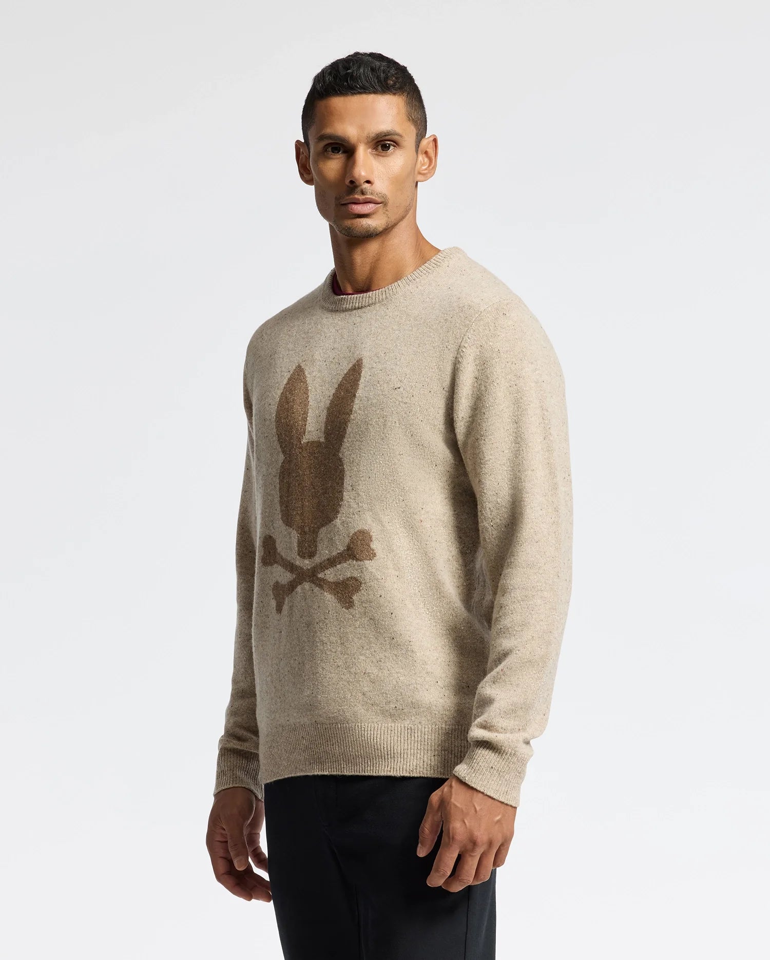 A man is pictured against a plain background donning the Psycho Bunny MENS LINCOLN MERINO WOOL SWEATER (B6E831E200) in beige, featuring a distinctive rabbit head with crossbones design. Crafted from cozy merino wool, it pairs seamlessly with his dark pants. His short hair complements the casual yet stylish essence of this look.