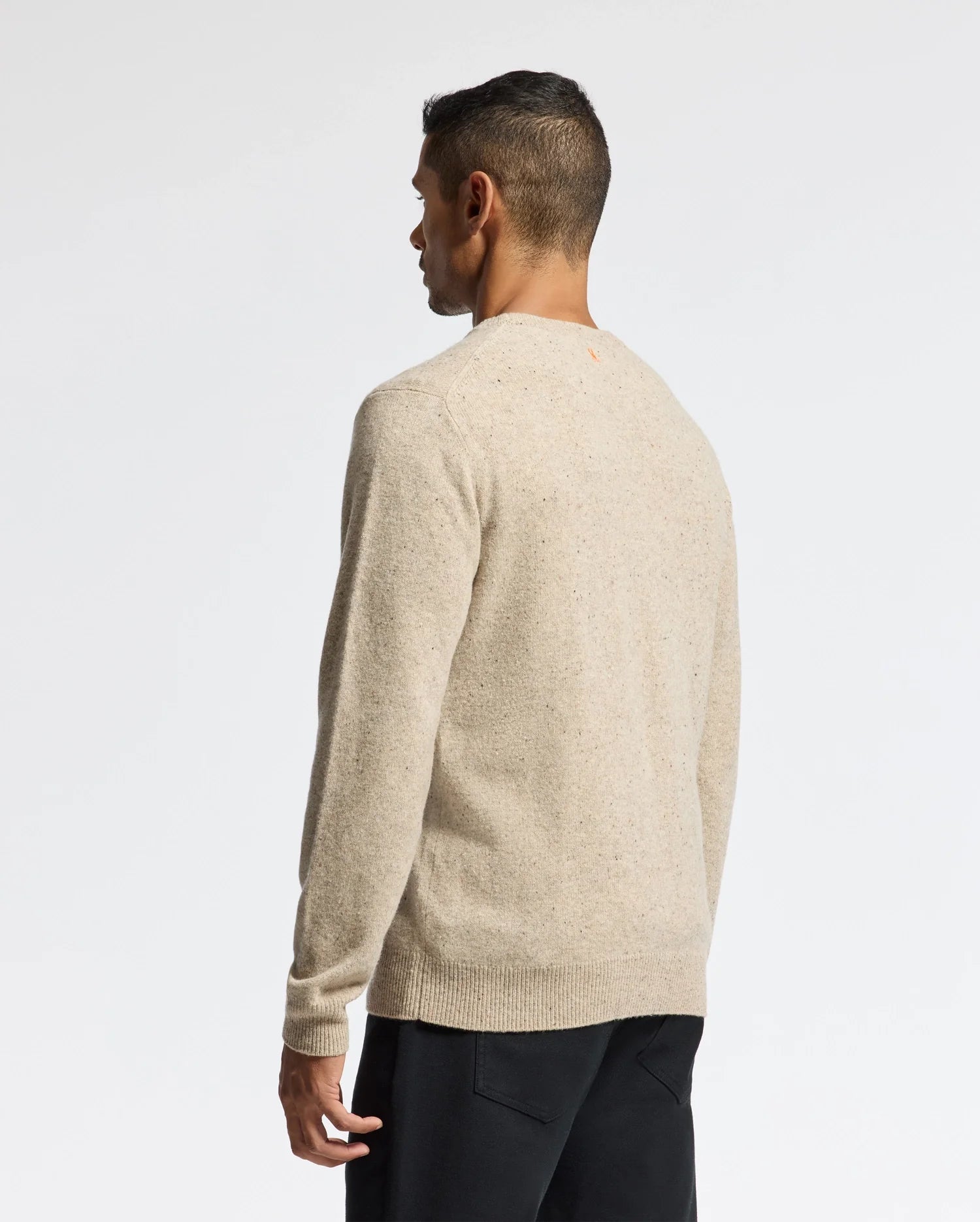 A man with short dark hair is facing away, wearing a beige MENS LINCOLN MERINO WOOL SWEATER (B6E831E200) by Psycho Bunny and black pants. The background is plain white.