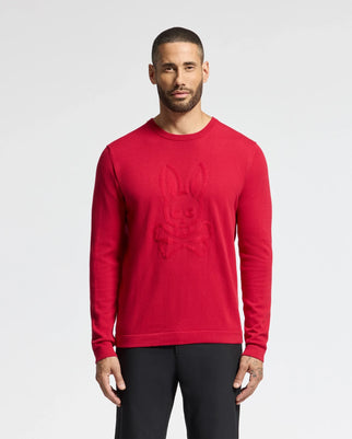 A person is wearing the Psycho Bunny Men's Cory Chainstitch Cotton Sweater, showcasing intricate embroidery of a bunny and crossbones on the front. Against a plain light backdrop, they complement it with sleek black pants for a stylish look.