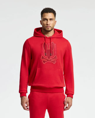 A man is strikingly dressed in a red cotton MENS PIERCE ESSENTIAL FRENCH TERRY HOODIE - B6H135E200 and matching sweatpants. The embroidered hoodie from Psycho Bunny showcases a stylized graphic of a bunny with crossbones, creating an edgy yet playful vibe. He stands confidently against the plain white background.