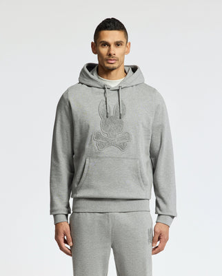 A man wearing the MENS ELDORADO BRUSH BACK HOODIE by Psycho Bunny in cozy grey brush-back cotton-blend stands against a white backdrop, showcasing the hoodie’s front design of a stylized bunny with crossed bones.