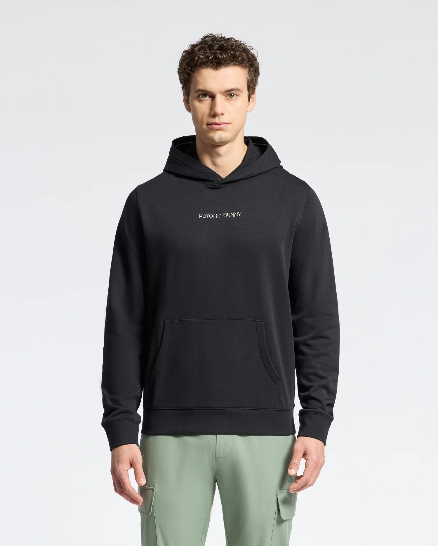 A young man with short curly hair wears a Psycho Bunny MENS NORA CLASSIC FRENCH TERRY WORDMARK HOODIE (B6H434F200) featuring a kangaroo pocket and chest logo, paired with light green pants, standing against a plain light background.