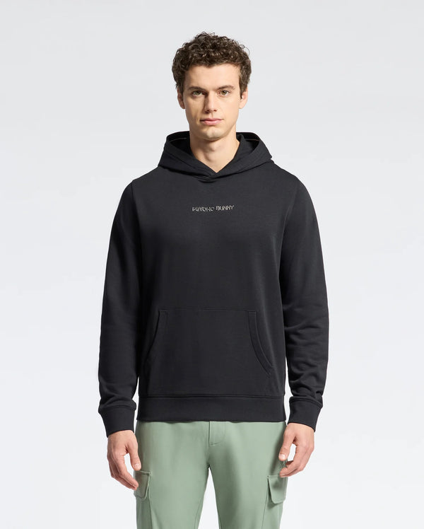 A young man with short curly hair wears a Psycho Bunny MENS NORA CLASSIC FRENCH TERRY WORDMARK HOODIE (B6H434F200) featuring a kangaroo pocket and chest logo, paired with light green pants, standing against a plain light background.