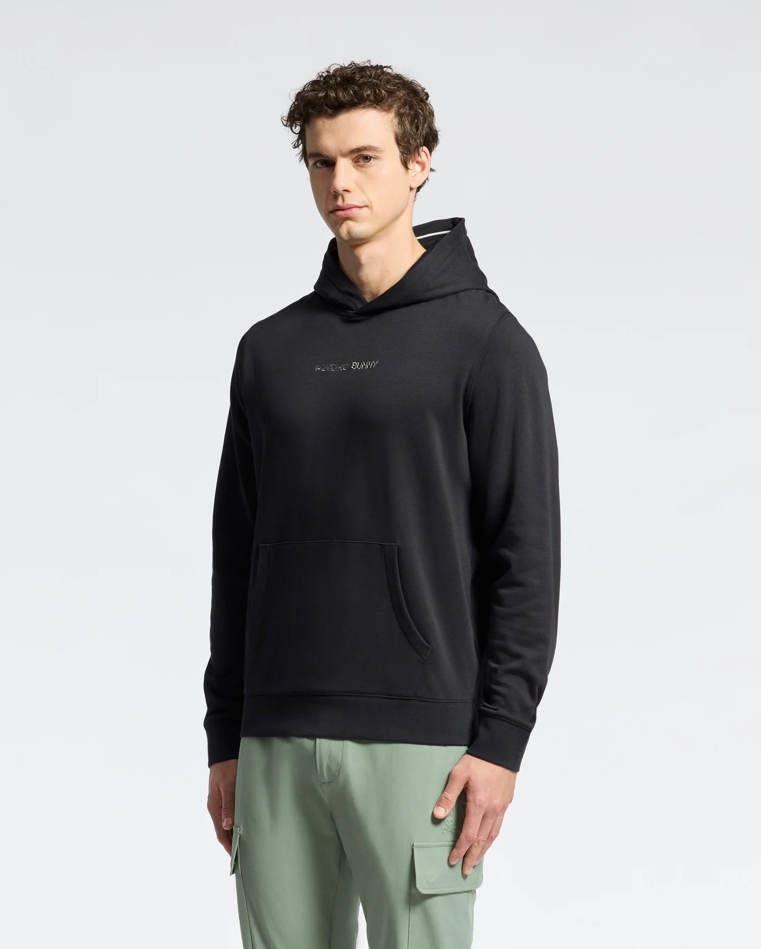 A man with short curly hair wears a black Psycho Bunny Mens Nora Classic French Terry Wordmark Hoodie with an embroidered chest logo and light green cargo pants, standing against a plain light background.