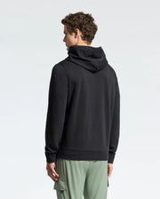 A person with short curly hair stands with their back to the camera, wearing a Psycho Bunny MENS NORA CLASSIC FRENCH TERRY WORDMARK HOODIE (B6H434F200) in black and light green cargo pants against a plain light background.