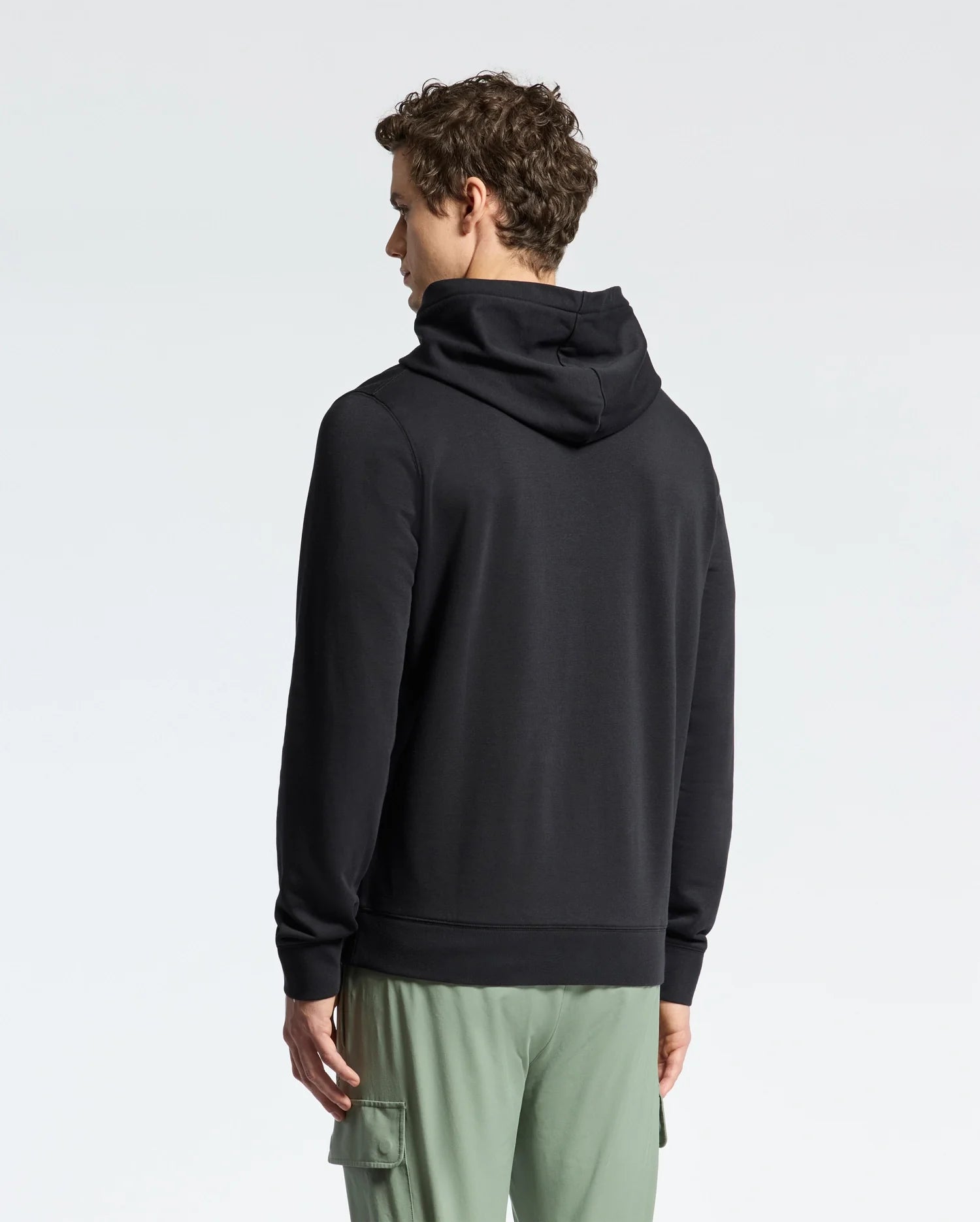 A person with short curly hair stands with their back to the camera, wearing a Psycho Bunny MENS NORA CLASSIC FRENCH TERRY WORDMARK HOODIE (B6H434F200) in black and light green cargo pants against a plain light background.