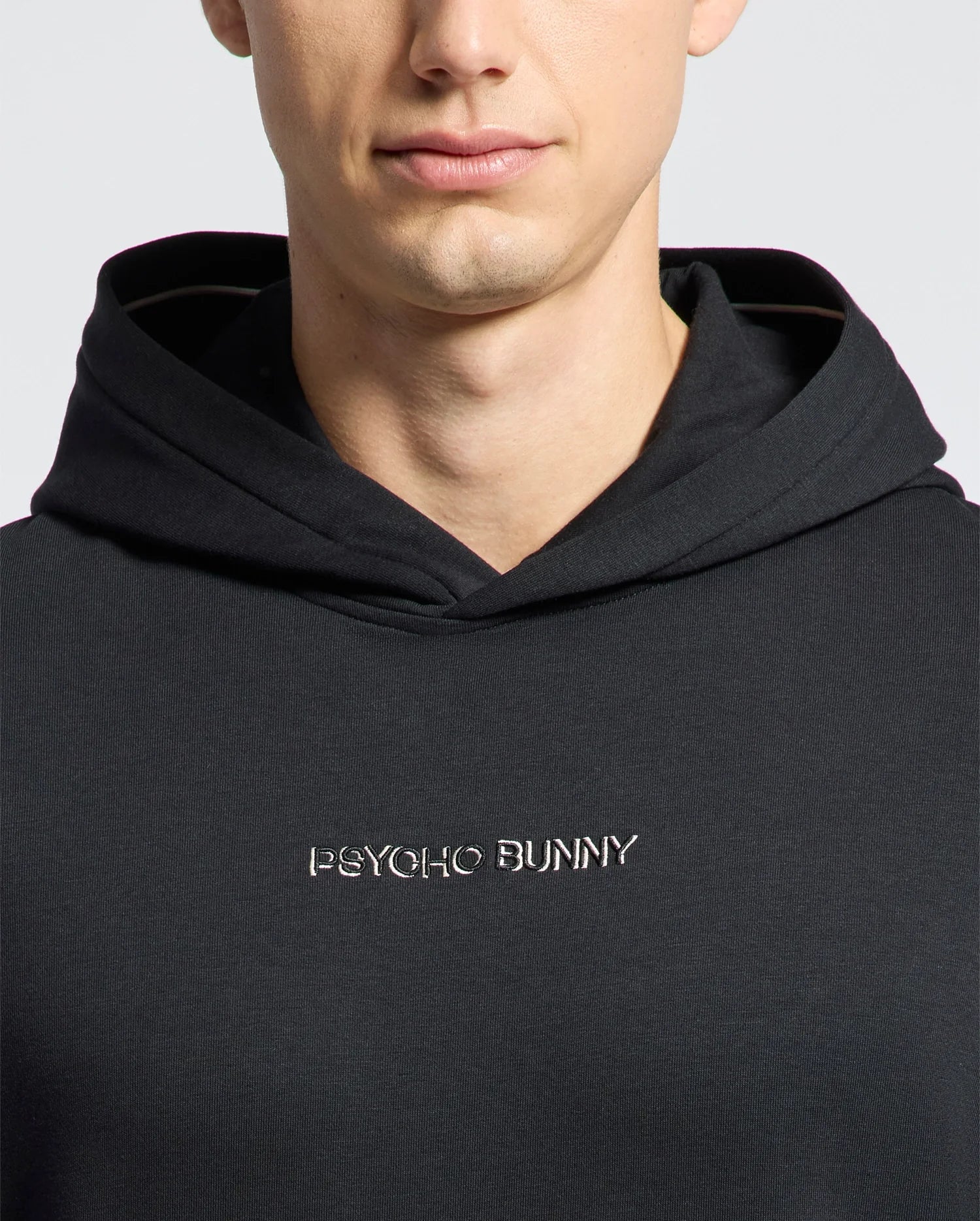 A person wears the Psycho Bunny Men's Nora Classic French Terry Wordmark Hoodie (B6H434F200) in black, with a bold embroidered logo on the front. Only the lower half of their face is visible.