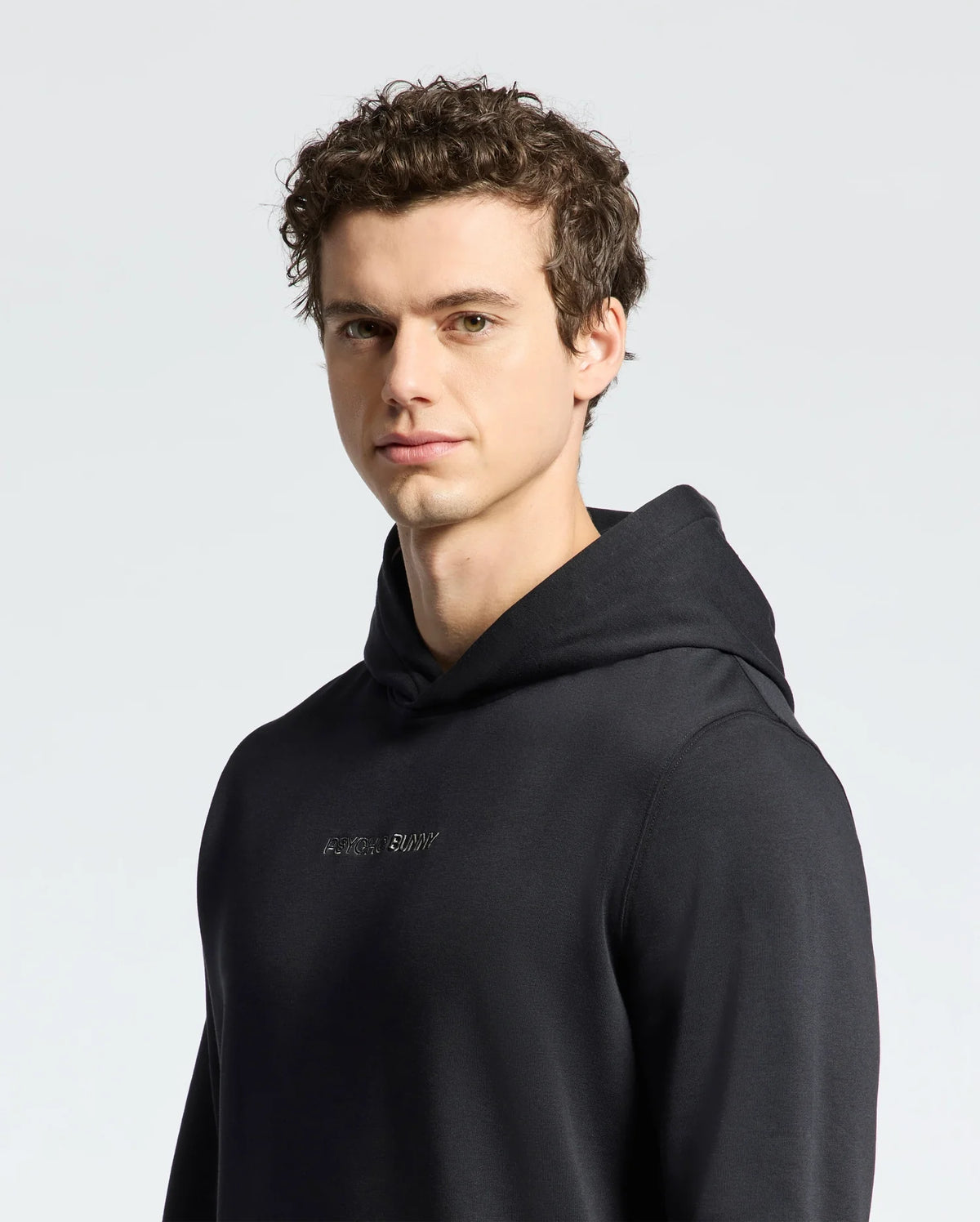 A young man with short, curly brown hair wears a Psycho Bunny MENS NORA CLASSIC FRENCH TERRY WORDMARK HOODIE (B6H434F200) in black, looking toward the camera against a plain light background.