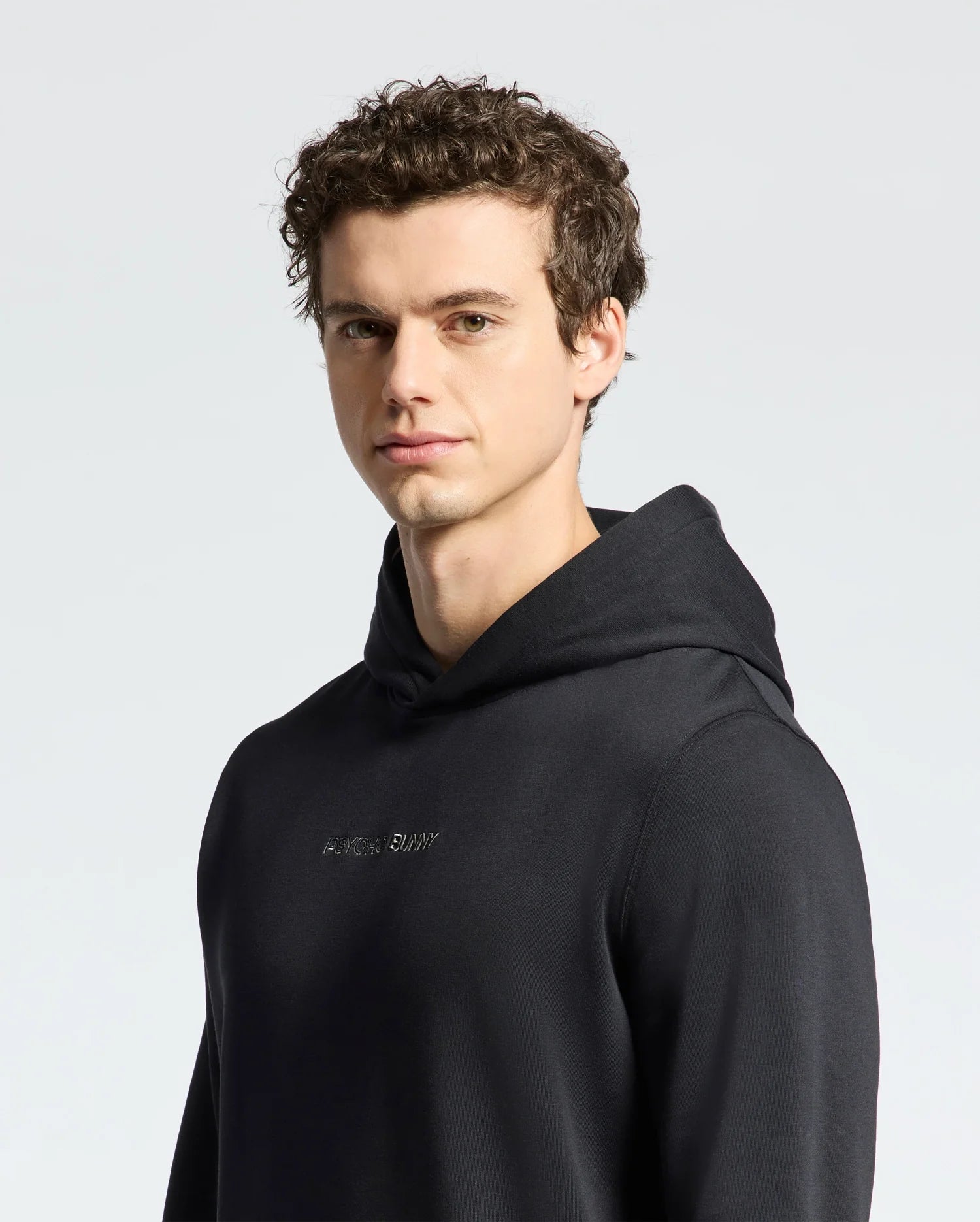 A young man with short, curly brown hair wears a Psycho Bunny MENS NORA CLASSIC FRENCH TERRY WORDMARK HOODIE (B6H434F200) in black, looking toward the camera against a plain light background.