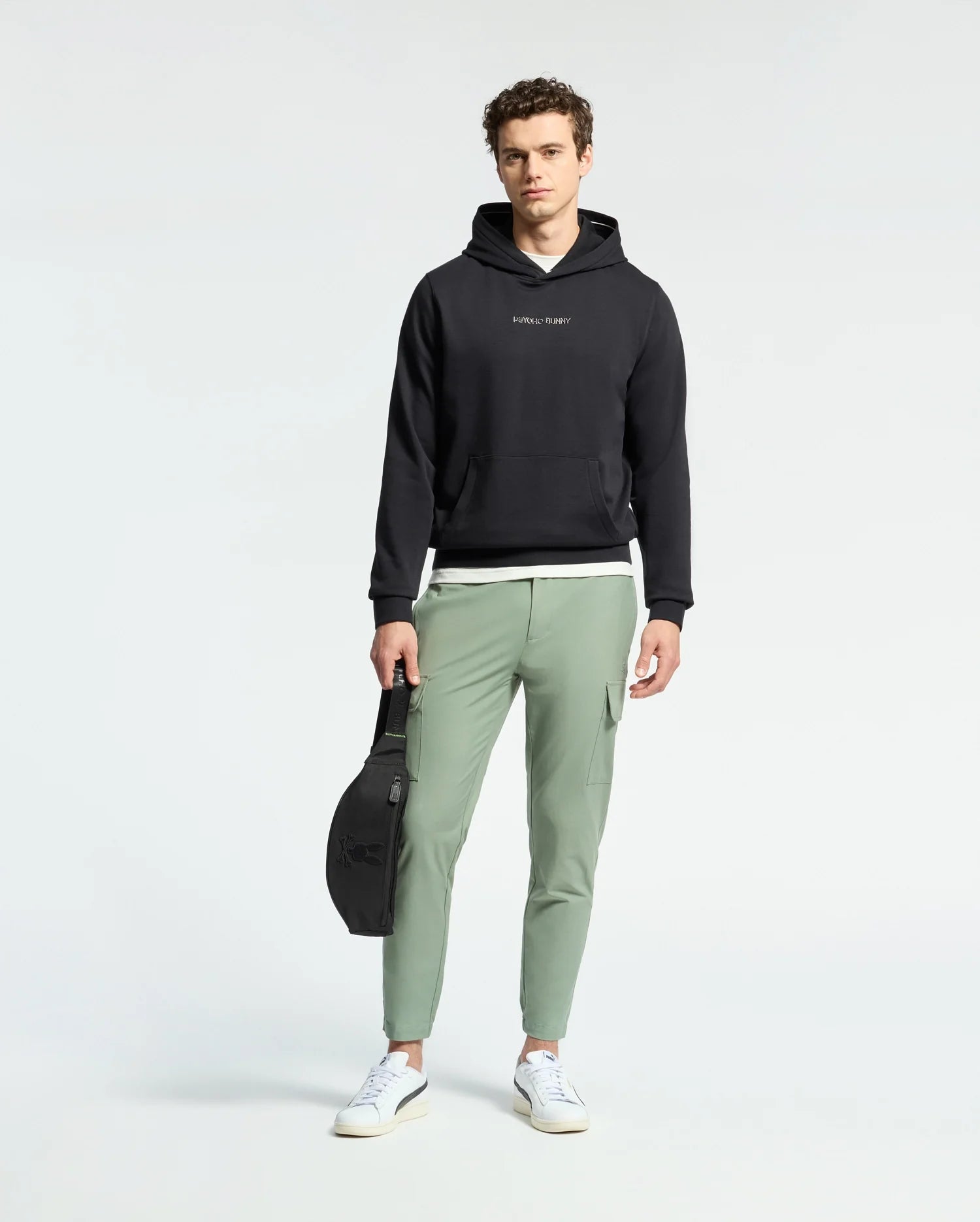 A man faces forward against a plain light background, wearing a black Psycho Bunny MENS NORA CLASSIC FRENCH TERRY WORDMARK HOODIE, light green joggers, white sneakers, and holding a black bag in his left hand.
