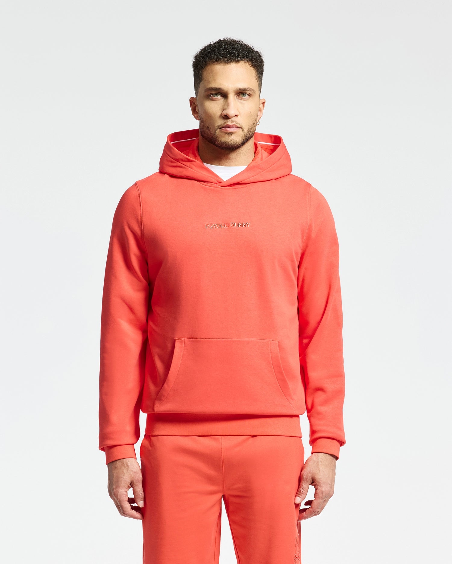 A man wears a matching red Psycho Bunny MENS NORA CLASSIC FRENCH TERRY WORDMARK HOODIE - B6H434F200 and pants. He stands against a plain white background, looking ahead. The hoodie includes a front pocket and wordmark embroidery across the chest.