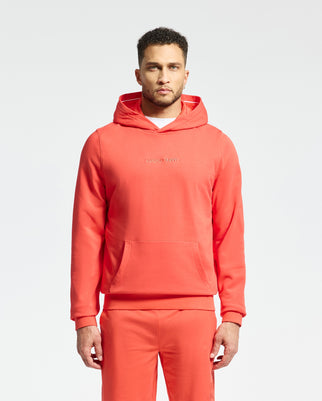 A man wears a matching red Psycho Bunny MENS NORA CLASSIC FRENCH TERRY WORDMARK HOODIE - B6H434F200 and pants. He stands against a plain white background, looking ahead. The hoodie includes a front pocket and wordmark embroidery across the chest.