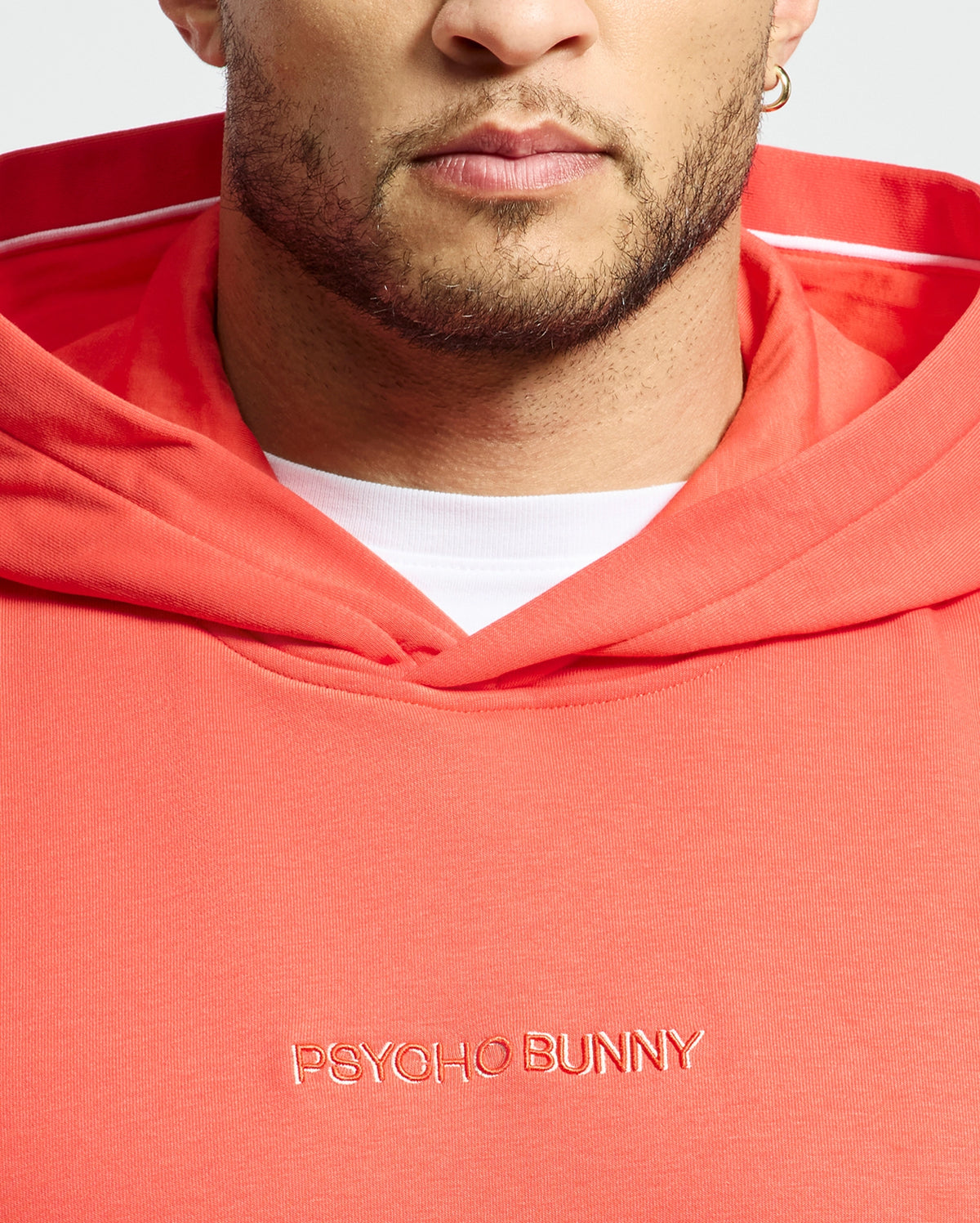 A person wears a coral MENS NORA CLASSIC FRENCH TERRY WORDMARK HOODIE by Psycho Bunny, with "Psycho Bunny" embroidery. The hood reveals a white lining, and their partially visible face shows a beard and earring.