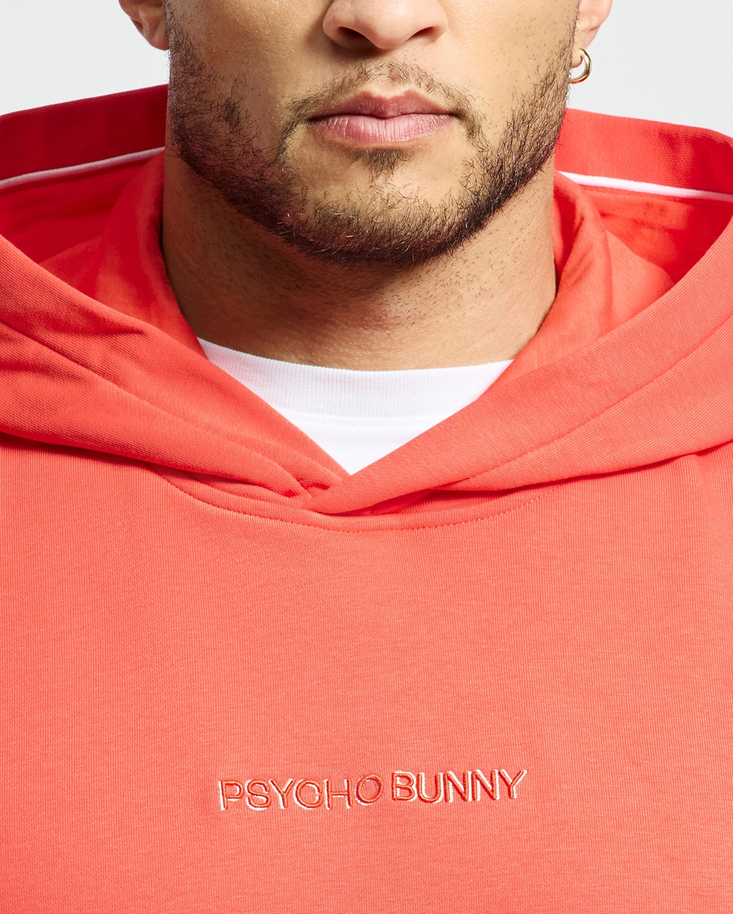 A person wears a coral MENS NORA CLASSIC FRENCH TERRY WORDMARK HOODIE by Psycho Bunny, with "Psycho Bunny" embroidery. The hood reveals a white lining, and their partially visible face shows a beard and earring.