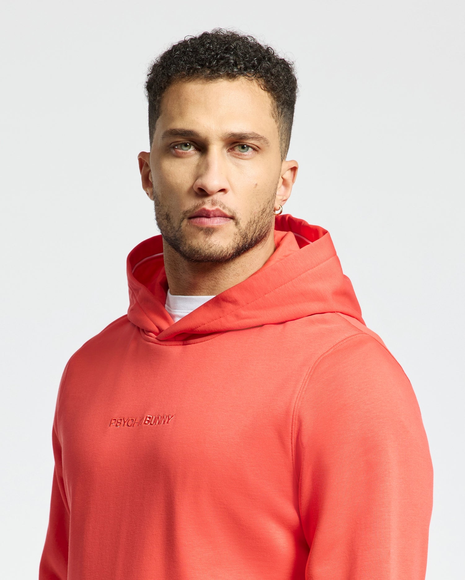 A man stands confidently against a white background, wearing the Men's Nora Classic French Terry Wordmark Hoodie - B6H434F200 by Psycho Bunny. The vibrant orange hoodie with "Psycho Bunny" embroidered boldly across the chest complements his short, curly hair and beard.