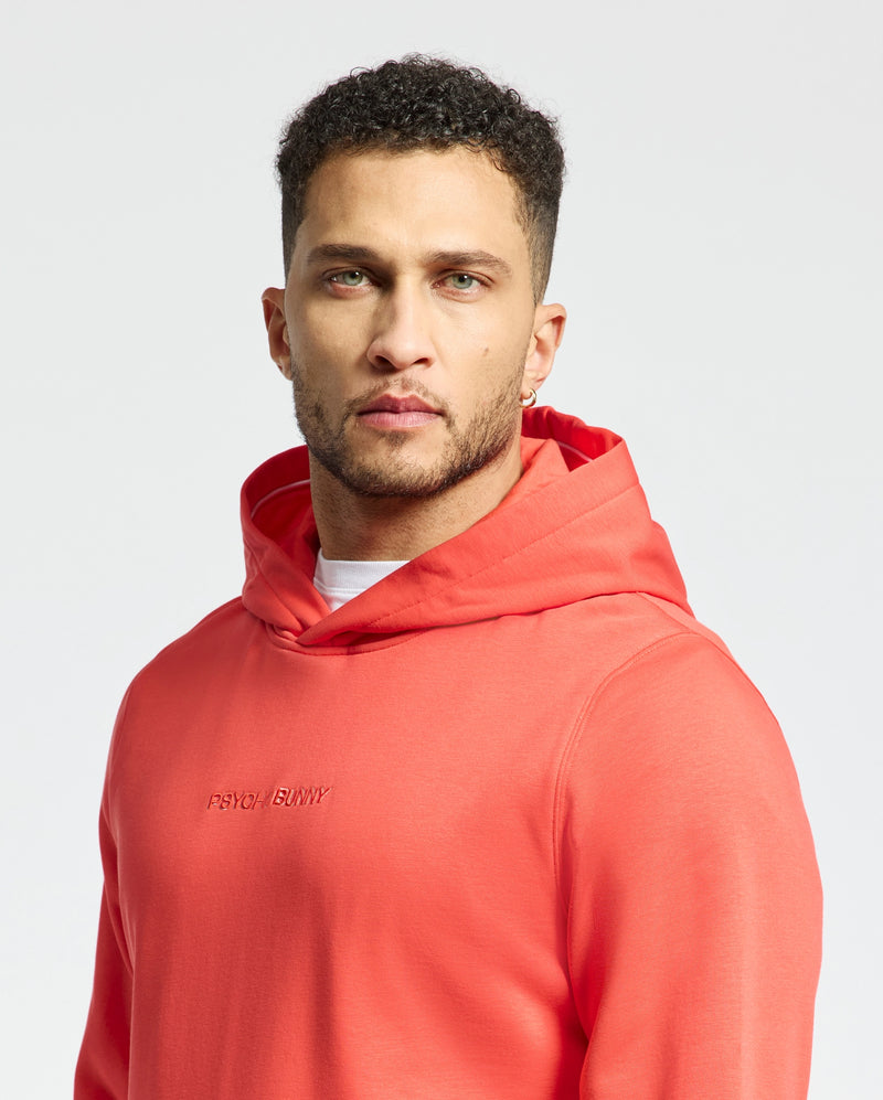A man stands confidently against a white background, wearing the Men's Nora Classic French Terry Wordmark Hoodie - B6H434F200 by Psycho Bunny. The vibrant orange hoodie with "Psycho Bunny" embroidered boldly across the chest complements his short, curly hair and beard.