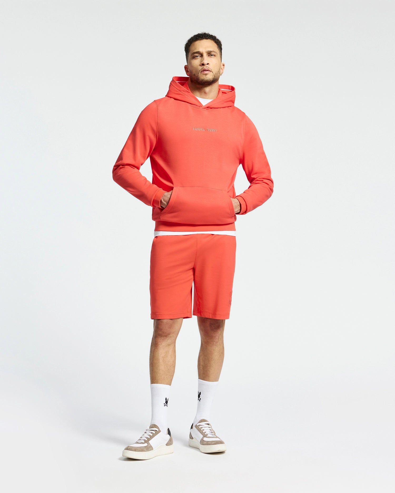 A man wears a matching coral ensemble, featuring the Psycho Bunny Nora Classic French Terry Wordmark Hoodie and shorts. His look is completed with white socks and beige sneakers against a plain off-white background.