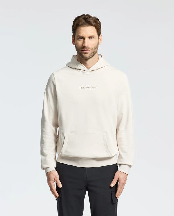 A man with short brown hair wears a light beige MENS NORA CLASSIC FRENCH TERRY WORDMARK HOODIE - B6H434F200 by Psycho Bunny, featuring an embroidered "Psycho Bunny" on the chest. Against a plain background, he pairs it with black pants and gazes at the camera neutrally.