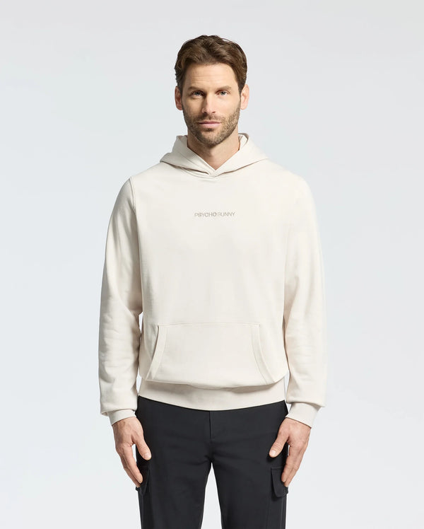 A man with short brown hair wears a light beige MENS NORA CLASSIC FRENCH TERRY WORDMARK HOODIE - B6H434F200 by Psycho Bunny, featuring an embroidered "Psycho Bunny" on the chest. Against a plain background, he pairs it with black pants and gazes at the camera neutrally.