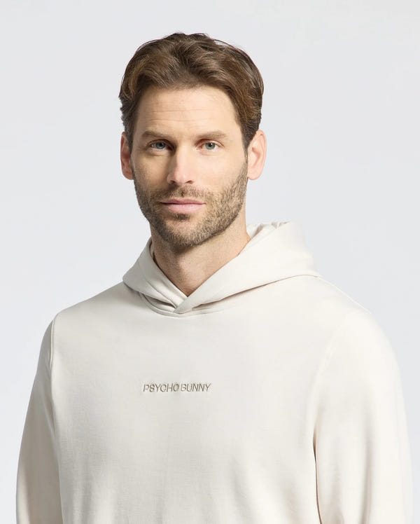 A man with short brown hair and a beard wears the MENS NORA CLASSIC FRENCH TERRY WORDMARK HOODIE by Psycho Bunny. This light-colored hoodie, made from soft French terry, features "Psycho Bunny" embroidered on the front and stands out against a plain background.