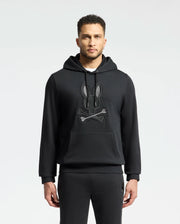 A man sports the Psycho Bunny MENS NEW CASTLE EMBROIDERED FRENCH TERRY HOODIE - B6H517F200, featuring a black bunny and crossbones design. With short hair, a trimmed beard, and matching pants, he embodies New Castle street style against a simple backdrop.