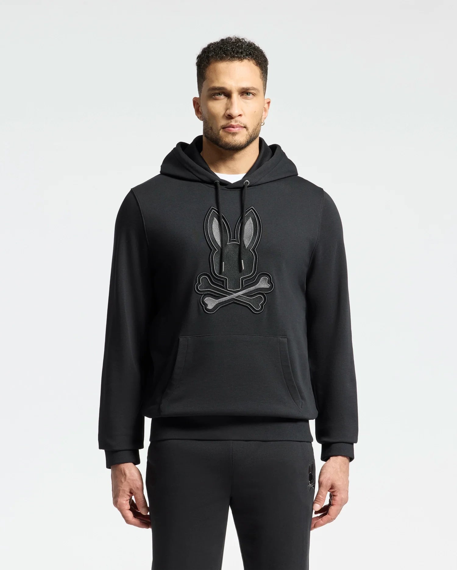 A man sports the Psycho Bunny MENS NEW CASTLE EMBROIDERED FRENCH TERRY HOODIE - B6H517F200, featuring a black bunny and crossbones design. With short hair, a trimmed beard, and matching pants, he embodies New Castle street style against a simple backdrop.