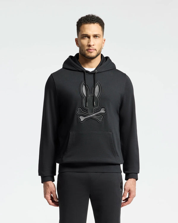 A man sports the Psycho Bunny MENS NEW CASTLE EMBROIDERED FRENCH TERRY HOODIE - B6H517F200, featuring a black bunny and crossbones design. With short hair, a trimmed beard, and matching pants, he embodies New Castle street style against a simple backdrop.