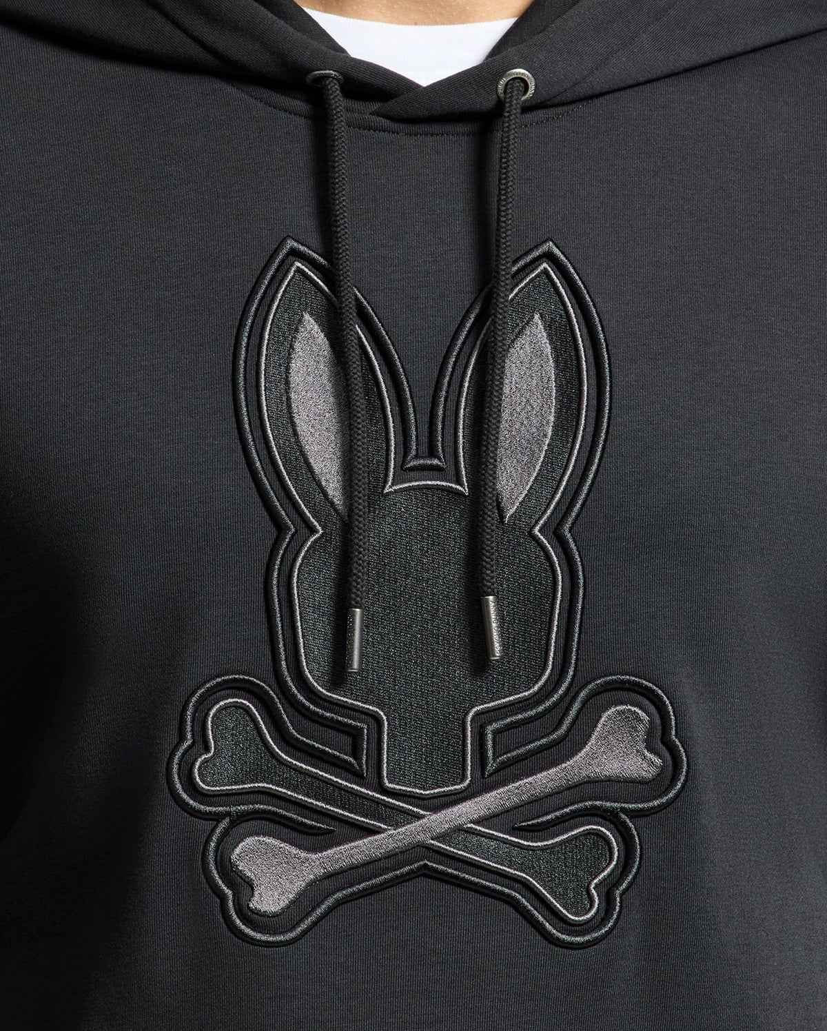 The Psycho Bunny Men's New Castle Embroidered French Terry Hoodie features a rabbit's head above crossed bones on black fabric. It includes black drawstrings, capturing New Castle's edgy style.