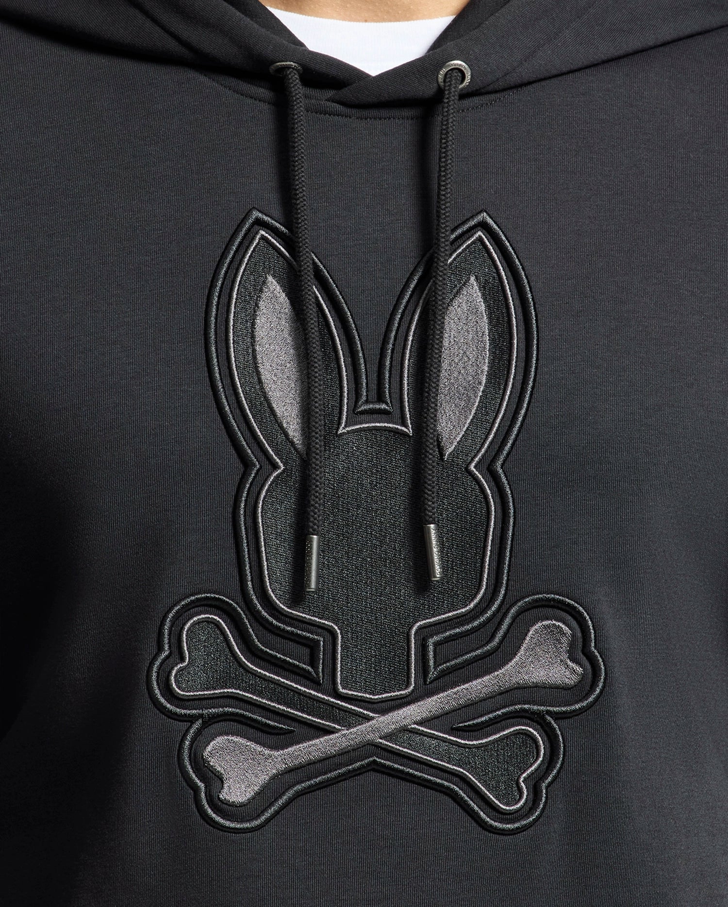 The Psycho Bunny Men's New Castle Embroidered French Terry Hoodie features a rabbit's head above crossed bones on black fabric. It includes black drawstrings, capturing New Castle's edgy style.