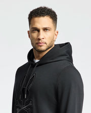 A person with short curly hair and an earring looks at the camera, wearing a Psycho Bunny Men's New Castle Embroidered French Terry Hoodie in black. The plain, pale background accentuates their presence in the foreground.