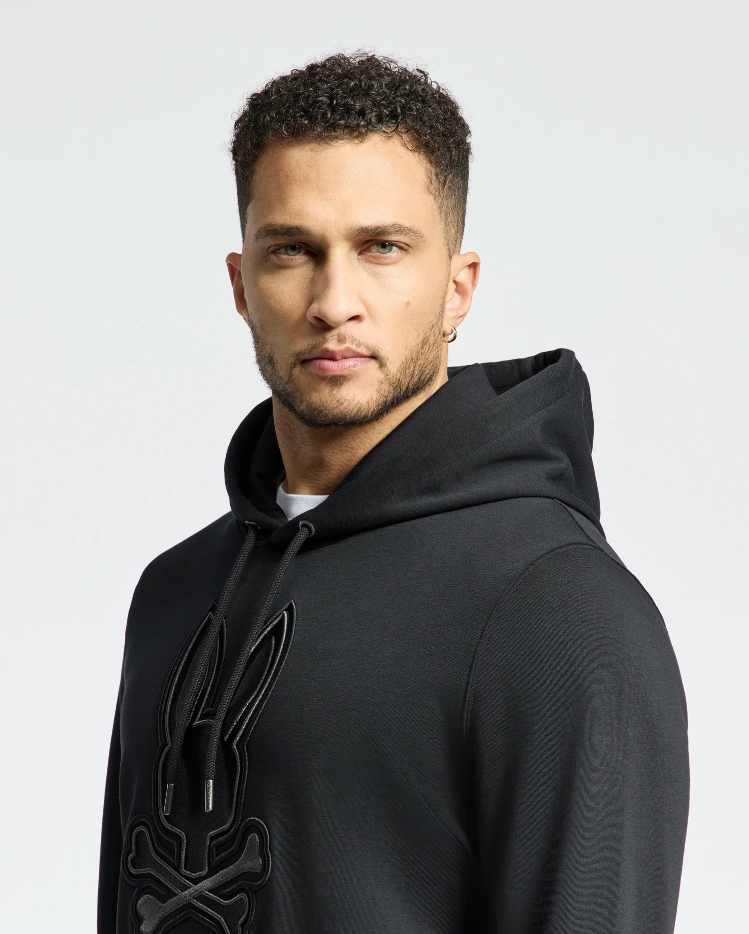 A person with short curly hair and an earring looks at the camera, wearing a Psycho Bunny Men's New Castle Embroidered French Terry Hoodie in black. The plain, pale background accentuates their presence in the foreground.