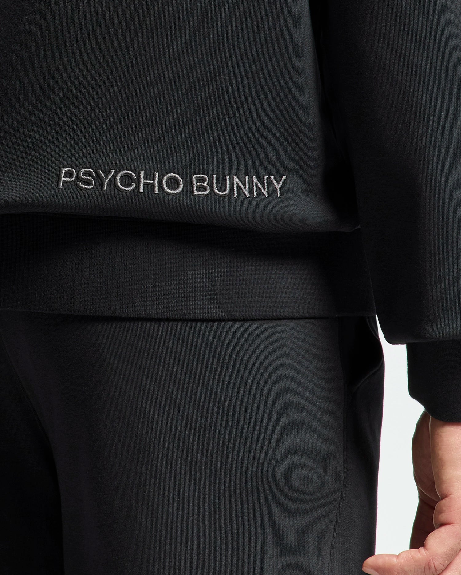 Close-up of someone in the Psycho Bunny MENS NEW CASTLE EMBROIDERED FRENCH TERRY HOODIE, with "Psycho Bunny" in white embroidery on the top's side; only part of the lower arm and upper body are visible, hinting at a style reflective of New Castle vibes.