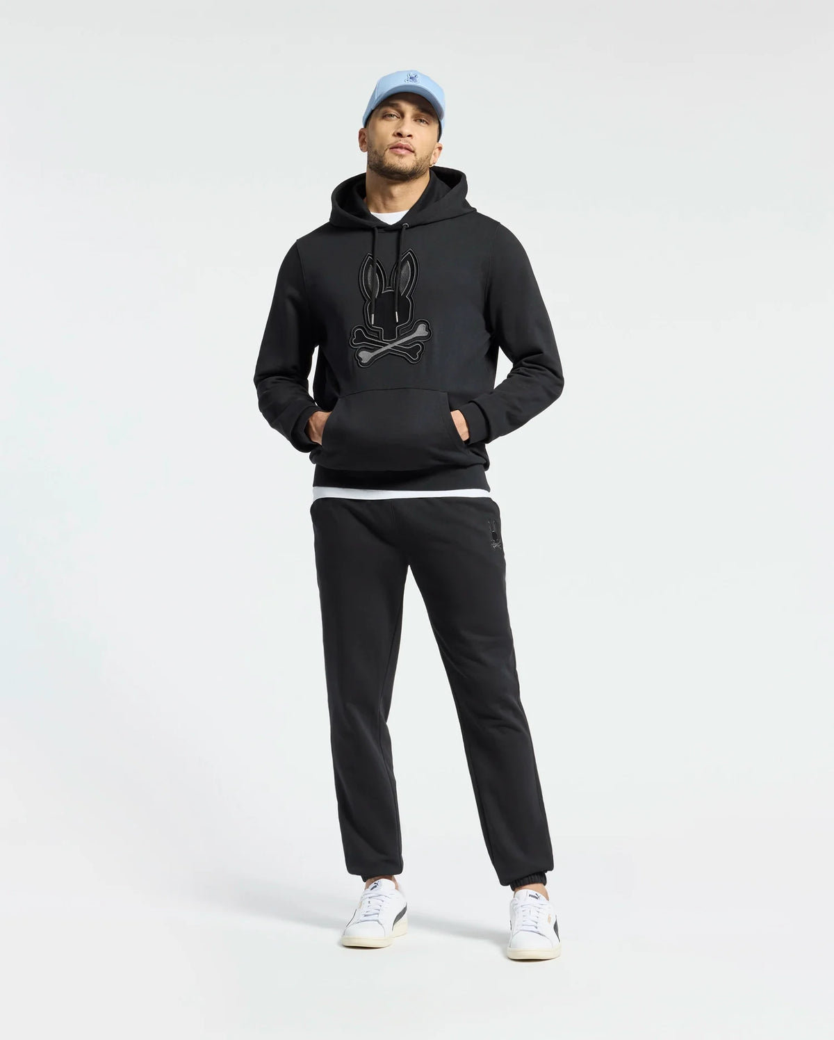 A man stands against a plain white background wearing a Psycho Bunny MENS NEW CASTLE EMBROIDERED FRENCH TERRY HOODIE in black, black joggers, a light blue cap, and white sneakers.