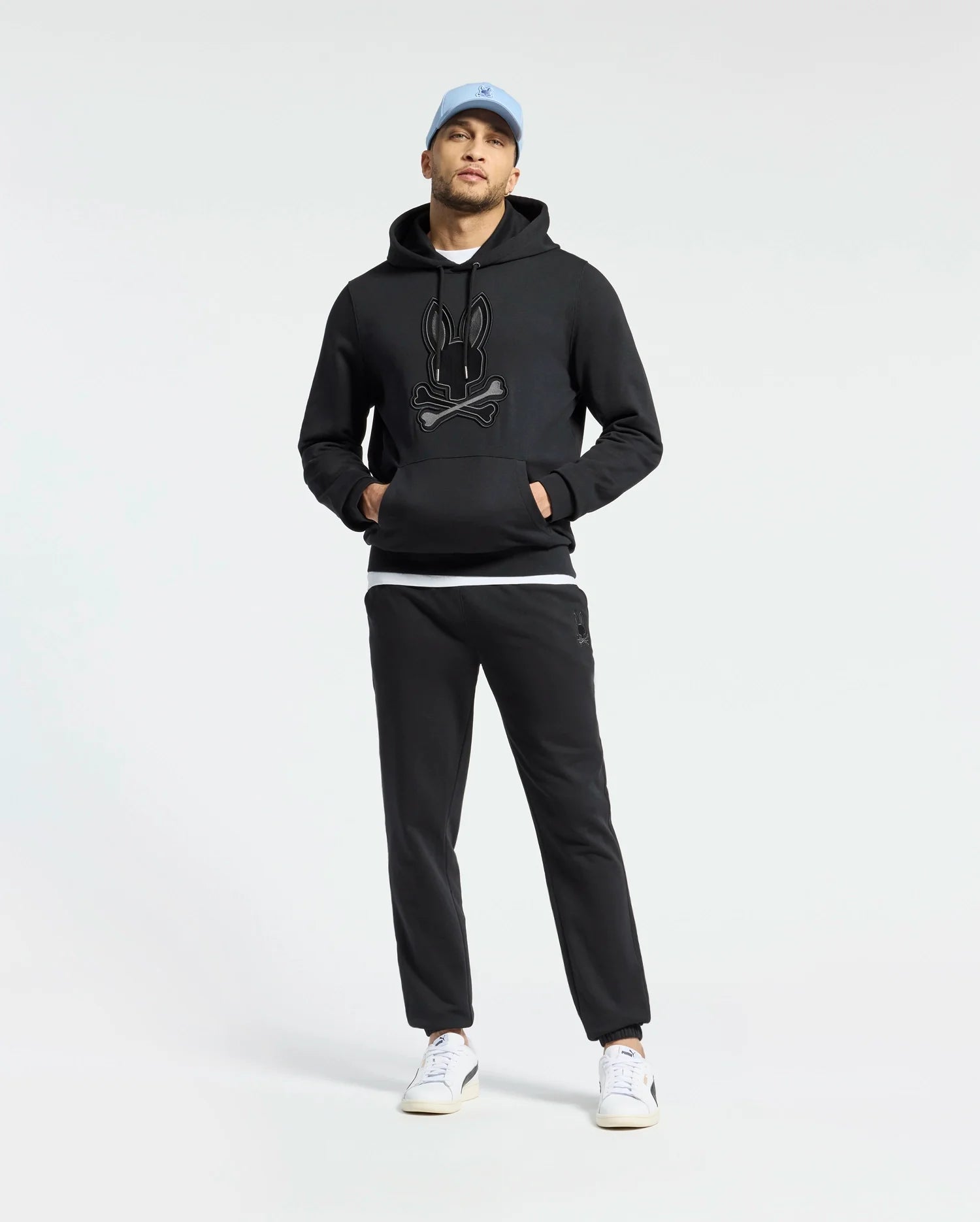 A man stands against a plain white background wearing a Psycho Bunny MENS NEW CASTLE EMBROIDERED FRENCH TERRY HOODIE in black, black joggers, a light blue cap, and white sneakers.