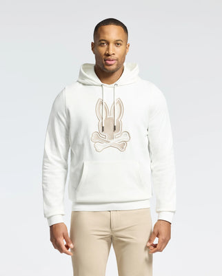 A man stands against a plain light background wearing a Psycho Bunny MENS NEW CASTLE EMBROIDERED FRENCH TERRY HOODIE, featuring a bunny and crossbones design. The white hoodie pairs seamlessly with his beige pants, while his short hair and relaxed posture add to the effortlessly elegant style.