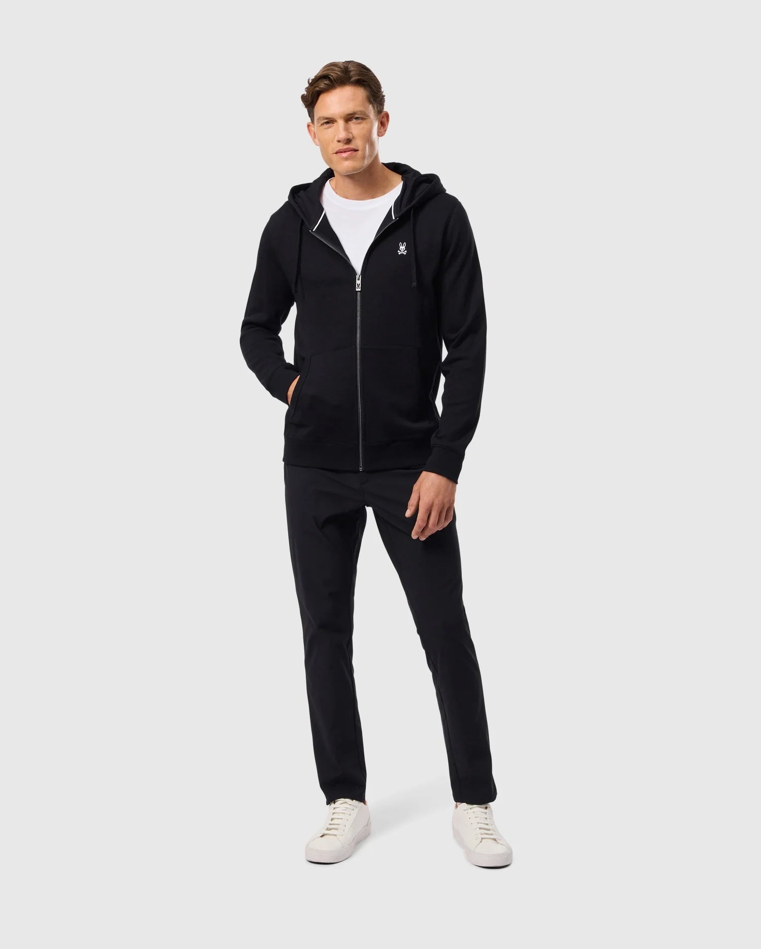 A person wearing a stylish Psycho Bunny MENS LEVY ZIP FLEECE HOODIE - B6H720D200 in black over a white T-shirt, paired with black pants and crisp white sneakers, stands against a plain light grey background.