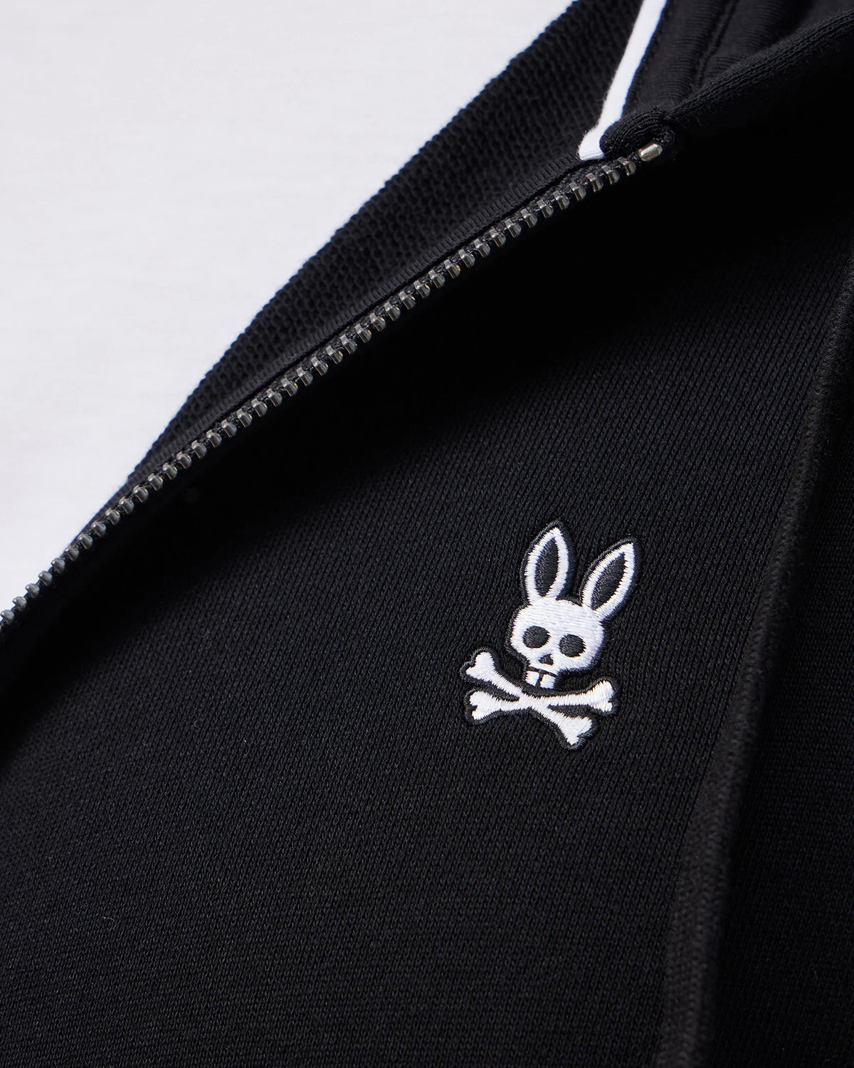 Close-up of the Psycho Bunny MENS LEVY ZIP FLEECE HOODIE - B6H720D200 in black, featuring a sleek white zipper and an embroidered cartoon rabbit skull and crossbones design on the fabric.
