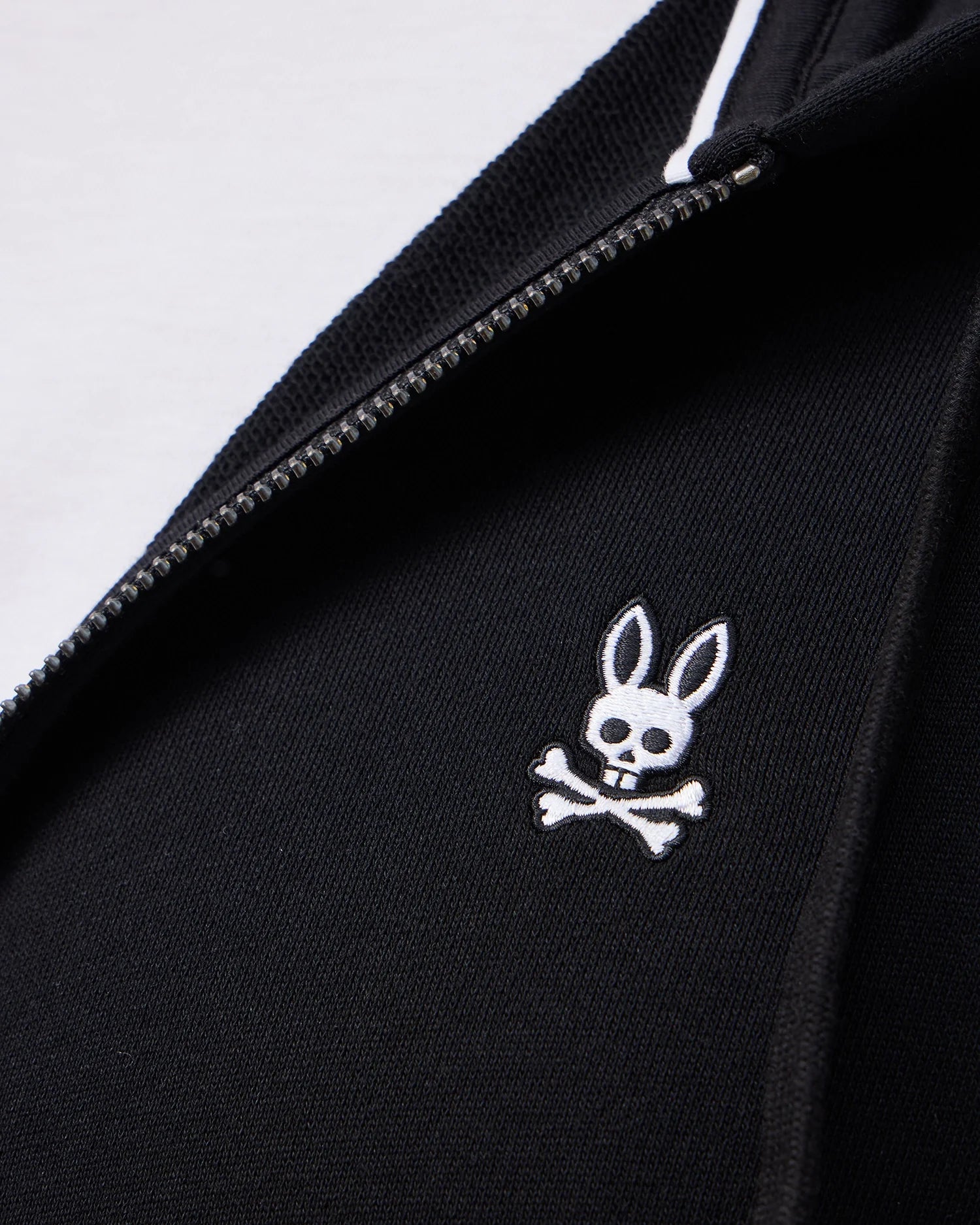 Close-up of the Psycho Bunny MENS LEVY ZIP FLEECE HOODIE - B6H720D200 in black, featuring a sleek white zipper and an embroidered cartoon rabbit skull and crossbones design on the fabric.
