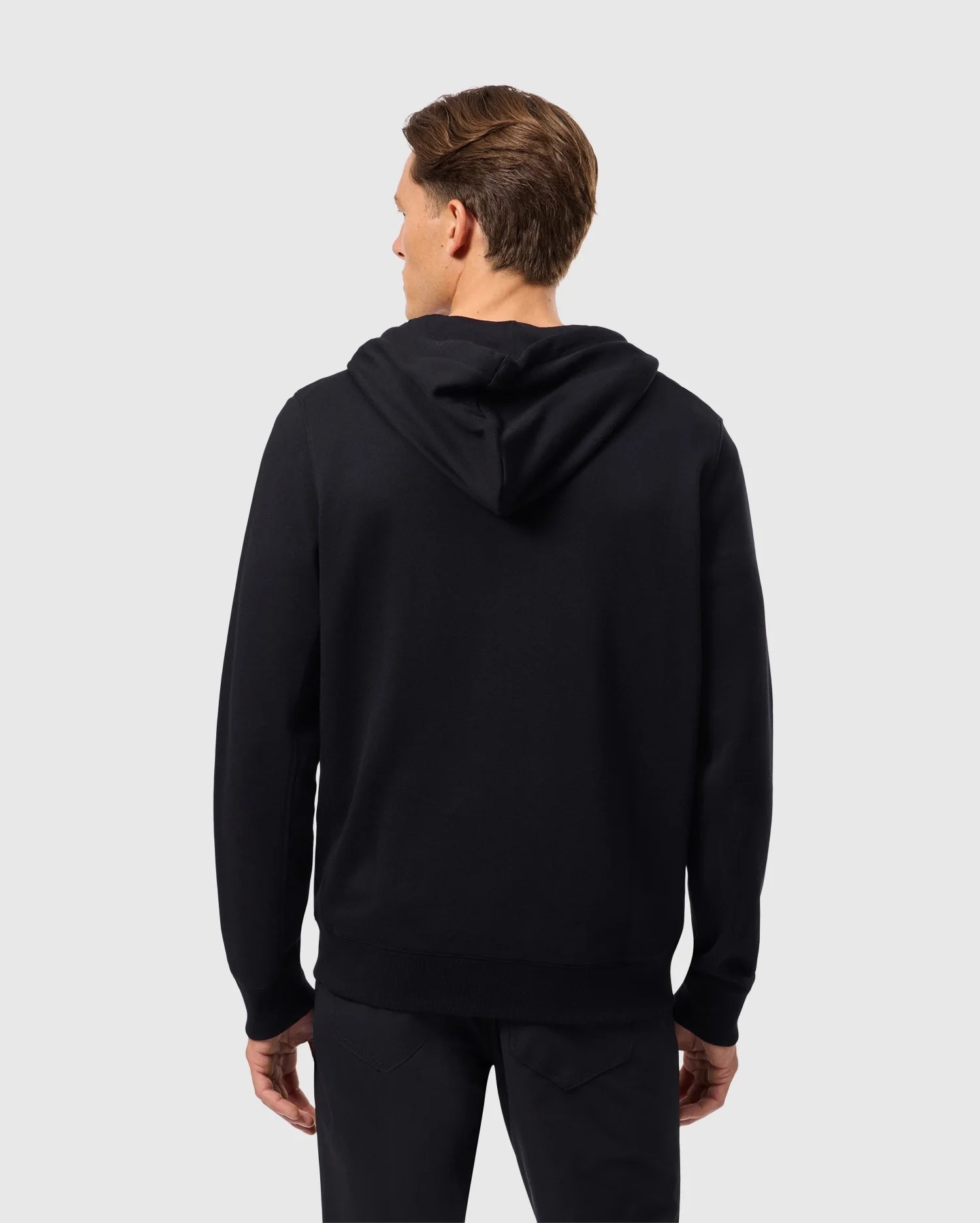 A person with short hair is wearing Psycho Bunny's Men's Levy Zip Fleece Hoodie (B6H720D200) and black pants, facing away from the camera. The background is plain gray.