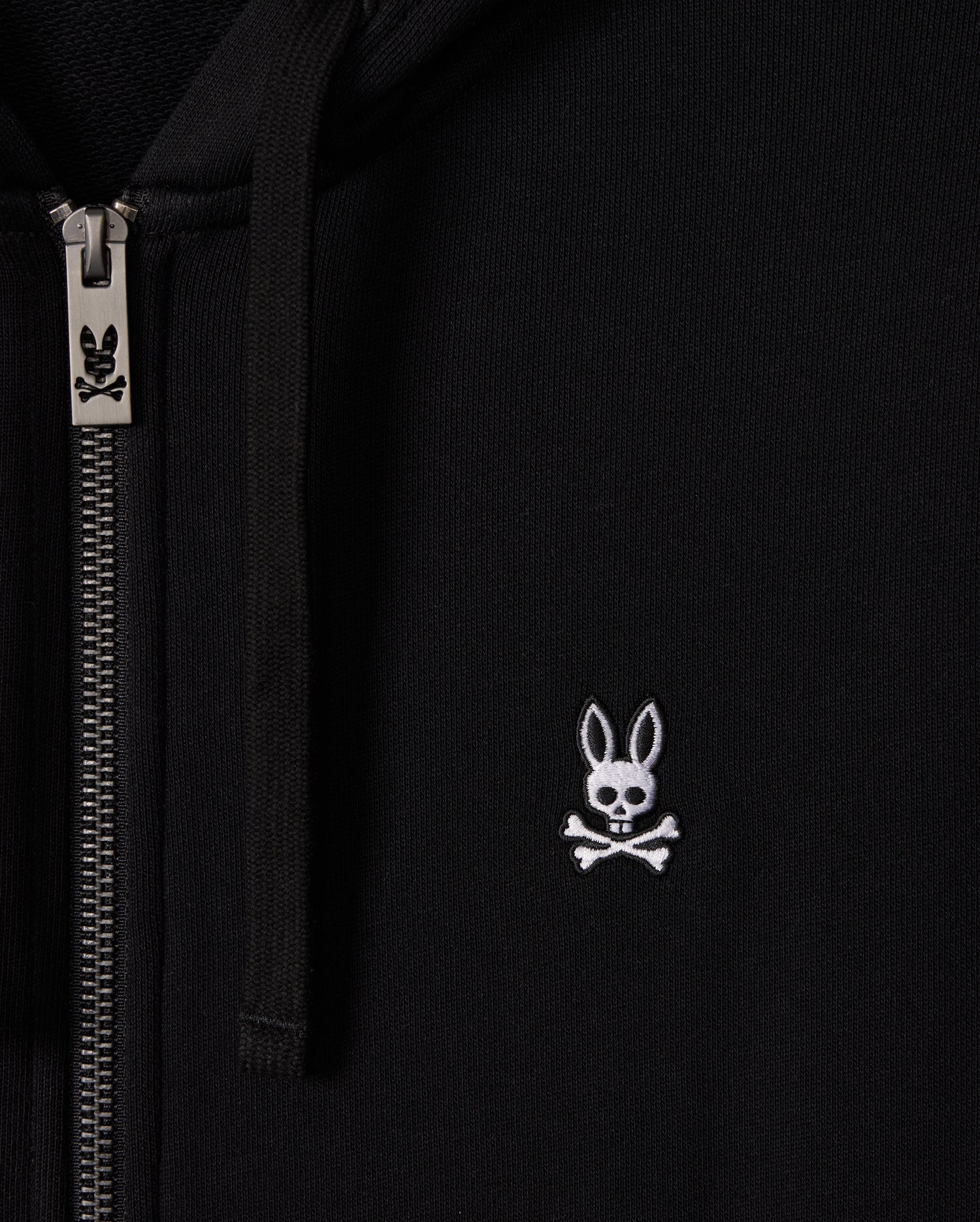 Close-up of the Psycho Bunny MENS LEVY ZIP FLEECE HOODIE - B6H720D200 in black, featuring a silver zipper on the left side. This cotton zip hoodie displays a small white logo of a rabbit skull and crossbones on the right side.
