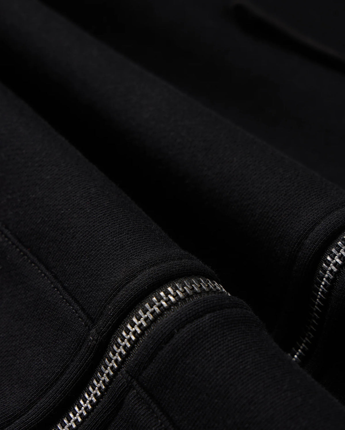 Here's a close-up shot of the MENS LEVY ZIP FLEECE HOODIE by Psycho Bunny, showcasing its black cotton fabric and two distinctive silver zippers. The contrast between the soft texture of the hoodie and the metallic sheen of the zippers highlights these intricate details on this men's zip hoodie.