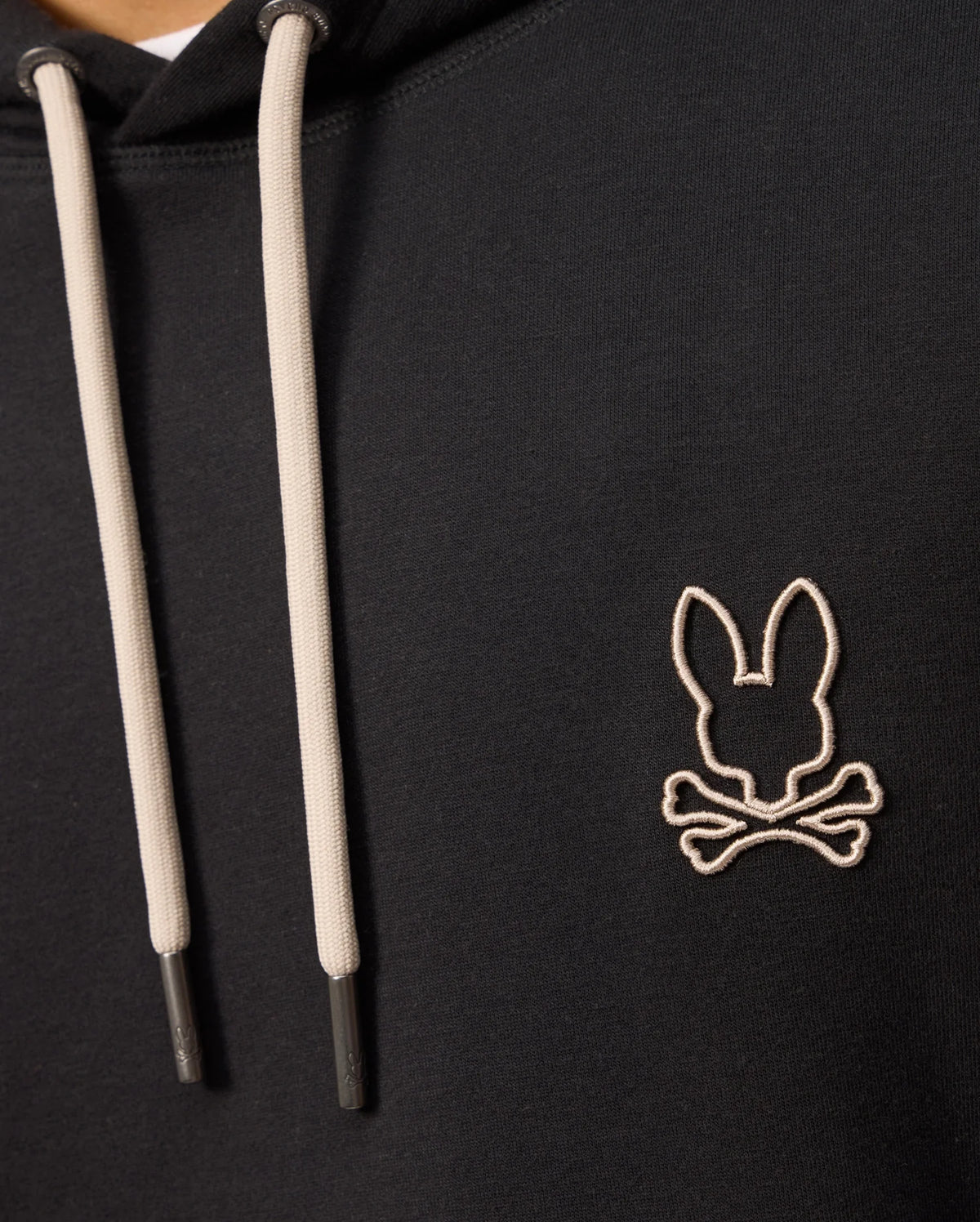 Close-up of the Psycho Bunny MENS JAMES PREMIUM HOODIE - B6H789D200 in black with light-colored drawstrings. This essential fleece hoodie showcases an embroidered bunny skull and crossbones on the chest. The image focuses on the upper part of the hoodie, revealing only part of the wearer's neck and shoulders.