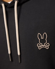Close-up of the Psycho Bunny MENS JAMES PREMIUM HOODIE - B6H789D200 in black with light-colored drawstrings. This essential fleece hoodie showcases an embroidered bunny skull and crossbones on the chest. The image focuses on the upper part of the hoodie, revealing only part of the wearer's neck and shoulders.