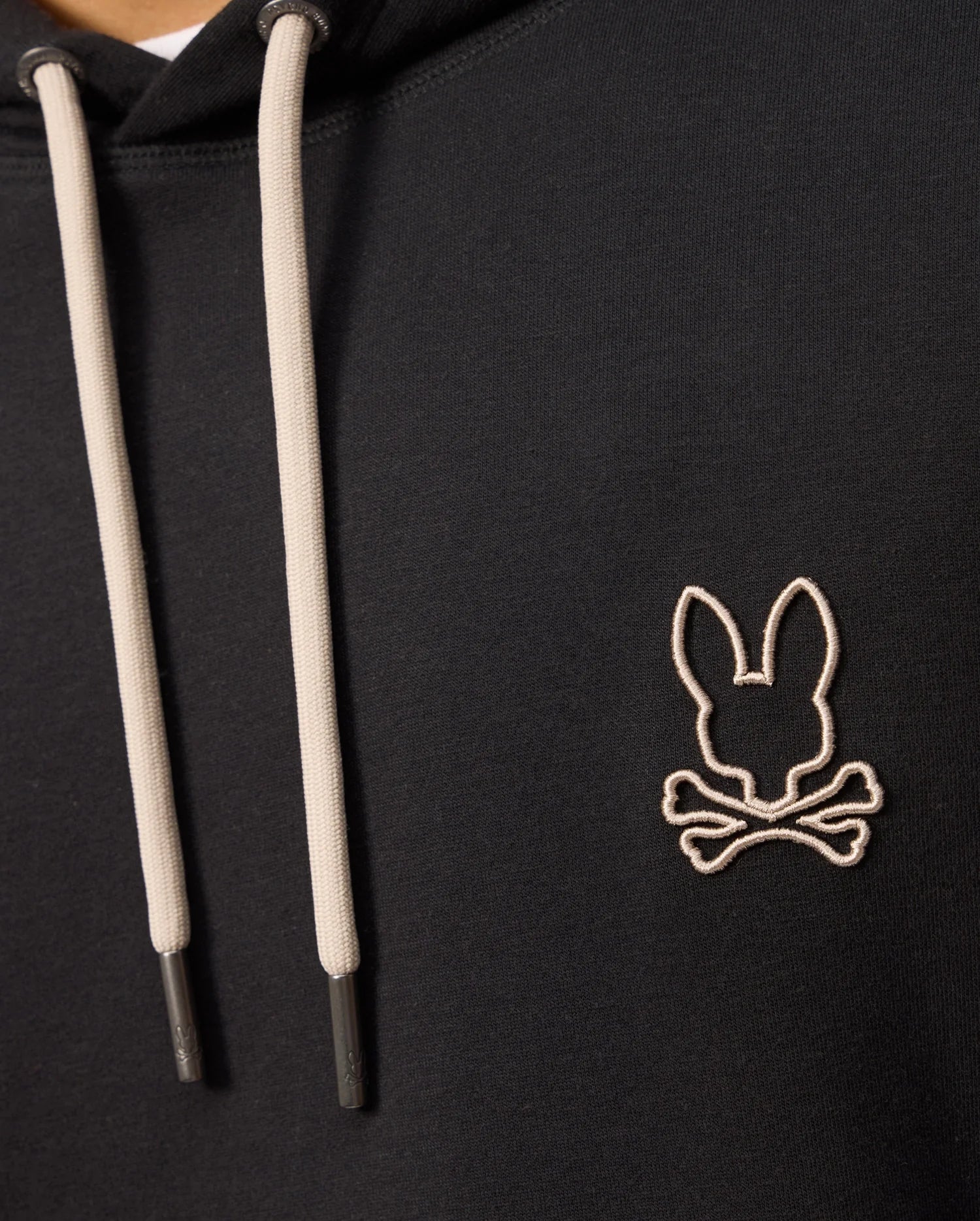 Close-up of the Psycho Bunny MENS JAMES PREMIUM HOODIE - B6H789D200 in black with light-colored drawstrings. This essential fleece hoodie showcases an embroidered bunny skull and crossbones on the chest. The image focuses on the upper part of the hoodie, revealing only part of the wearer's neck and shoulders.