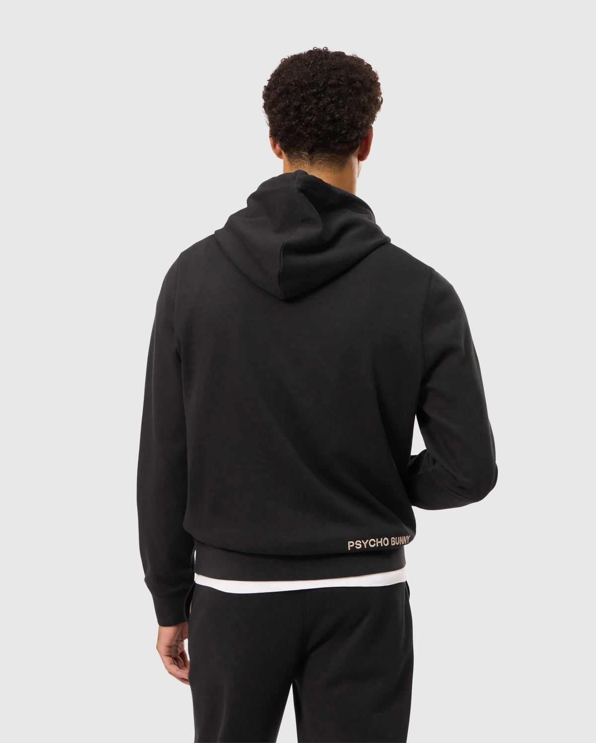 A person with short curly hair is shown from the back, wearing a black hoodie and black pants. The MENS JAMES PREMIUM HOODIE - B6H789D200 from Psycho Bunny features an embroidered bunny logo near the lower back that reads "Psycho Bunny." The background is plain light gray.