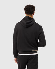A person with short curly hair is shown from the back, wearing a black hoodie and black pants. The MENS JAMES PREMIUM HOODIE - B6H789D200 from Psycho Bunny features an embroidered bunny logo near the lower back that reads "Psycho Bunny." The background is plain light gray.